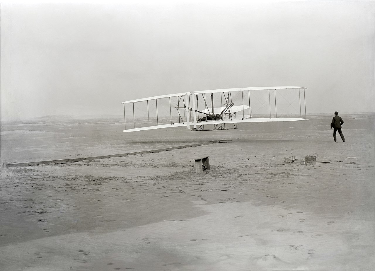 Restored image taken by John T. Daniels on December 17, 1903 near Kitty Hawk, North Carolina only seconds into the 12 second first powered flight piloted by Orville Wright. Wilbur Wright is seen to the right. Public Domain.